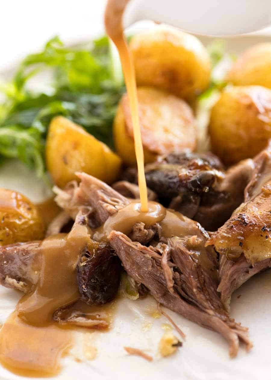 Gravy being poured over Slow Cooked Lamb Shoulder on a plate with a side of roast potatoes and salad, ready to be eaten
